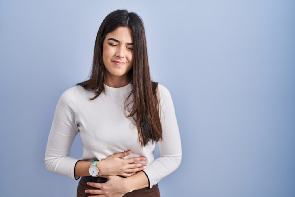 young brunette woman standing blue background with hand stomach because indigestion painful illness feeling unwell ache concept